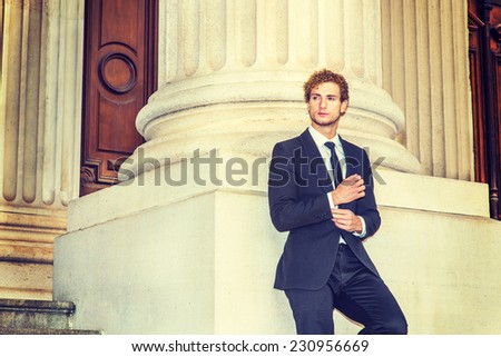 Man Looking for Success. Dressing in black suit with Shawl Lapel, black necktie, a young sexy guy with curly hair is standing by column outside office, a hand touching a cuff of sleeve, thinking.
