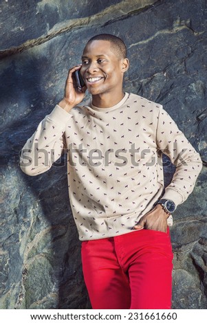 Man Talking on Phone. Dressing in cream, patterned, collarless sweater, red pants, wearing wristwatch, a young black guy is standing by rocks, smiling, making a phone call on his mobile phone.