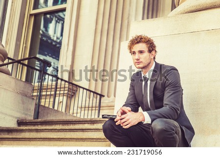 Man Waiting for You. Dressing in black suit with Shawl Lapel, black necktie, a young sexy guy with curly hair is squatting outside office, holding a laptop computer, relaxing.
