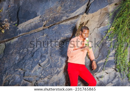 Man Missing You. Dressing in light orange sweater with high collar, red pants, wearing wristwatch, a young black guy is standing against rocks with long leaves, holding white rose, smelling, thinking.