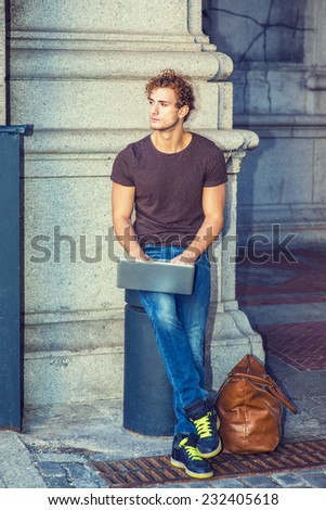 Wearing dark brown T shirt, blue jeans, black sneakers, leather bag on ground, a young sexy guy with curly hair is sitting on a metal stake on the corner, working on laptop computer, thinking.