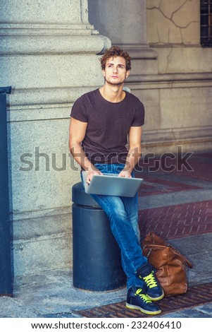 Wearing dark brown T shirt, jeans, sneakers, leather bag on the ground, a young sexy guy with curly hair is sitting on a metal stake on the street, looking up, working on laptop computer, thinking.