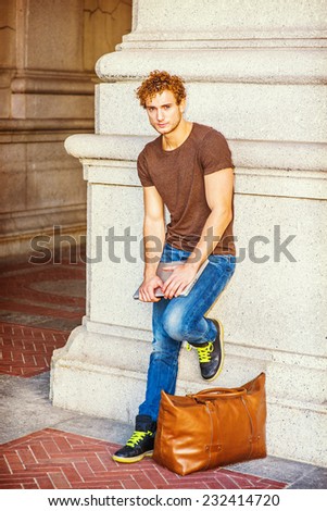 Traveler Relaxing on Street. Wearing dark brown T shirt, jeans, sneakers, a leather bag on ground. a young sexy guy with curly hair is standing against wall, holding laptop computer, looking at you.