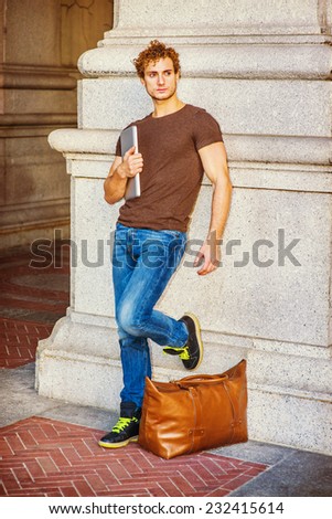 Traveler Relaxing on Street. Wearing dark brown T shirt, jeans, sneakers, a leather bag on ground. a young sexy guy with curly hair is standing against wall, holding laptop computer, waiting for you.