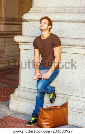 Man Relaxing on Street. Wearing dark brown T shirt, jeans, sneakers, a leather bag on ground. a young sexy guy with curly hair is standing against wall, holding laptop computer, looking up, thinking.