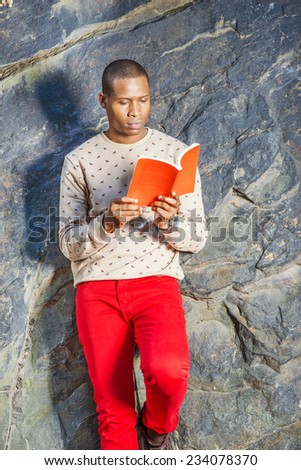 Man Reading Outside. Dressing in cream, patterned, collarless sweater, red jeans, wearing wristwatch, holding a red book, a young black guy is leaning against rocks, looking down, reading.