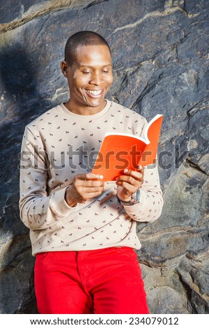 Man Reading Outside. Dressing in cream, patterned, collarless sweater, red jeans, wearing wristwatch, holding a red book, a young black guy is leaning against rocks, smiling, looking down, reading.