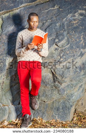 Man Reading Outside. Dressing in cream, patterned, collarless sweater, red jeans, boot shoes, wearing wristwatch, holding a red book, a young black guy is leaning against rocks, looking down, reading.