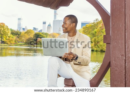 Business Man Working. Dressing in cream blazer, collarless sweater, white pants, wearing a wristwatch, a young black guy is sitting by lake in big city, smiling, thinking, working on laptop computer.