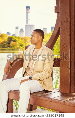 Man Waiting for You. Dressing in cream blazer, collarless sweater, white pants, wearing a wristwatch, a young black guy is sitting by a lake in a big city, relaxing, thinking.
