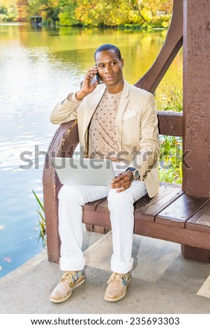Business Man Working Outside. Dressing in cream blazer, collarless sweater, white pants, sneakers, a young black guy is sitting by a lake, talking on phone, looking away, working on laptop computer.