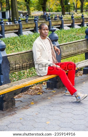Man Waiting for You. Dressing in cream blazer, collarless sweater, red pants, sneakers, a young black  guy is sitting on the bench on the park, looking at you, relaxing. Man Casual Fashion
