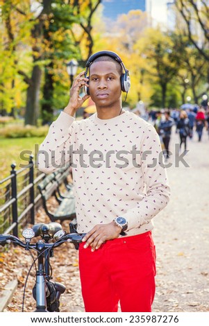 Man Listening Music. Dressing in collarless sweater, red pants, wearing headphone, a young black guy is standing on street, holding a bike, a hand touching headphone pad, seriously looking at you.