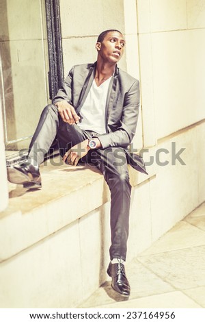 Young black man thinking outside. Wearing white under wear, fashionable jacket, pants, shoes, a young black student is sitting against window frame, sad, emotionally looking up. Street Fashion.