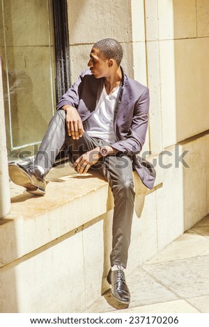Young black man thinking outside. Wearing a white under wear, fashionable jacket, pants, shoes, a young black college student is sitting against a window frame, sad, looking away. Street Fashion.