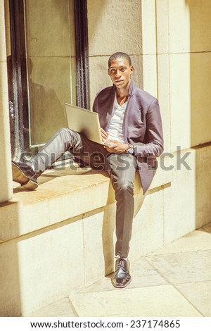 Man Studying Outside. Dressing in fashionable jacket, pants, leather shoes, wearing wristwatch, a young black student is sitting against window frame, reading, thinking, working on a laptop computer.
