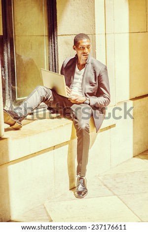 Man Working Outside. Dressing in fashionable jacket, pants, leather shoes, wearing wristwatch, a young black student is sitting against a window frame, reading, thinking, working on a laptop computer.