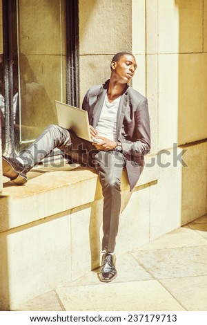 Difficult Task. Dressing in fashionable jacket, pants, leather shoes, a young black college student is sitting against a window frame, looking away, sad, thinking, working on a laptop computer.