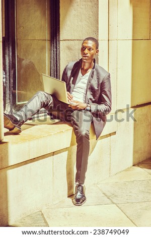 Man Studying Outside. Dressing in fashionable jacket, pants, leather shoes, wearing wristwatch, a young black student is sitting against a window frame, reading, thinking, working on laptop computer.