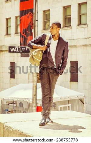 Street Fashion. Wearing a white under wear, fashionable jacket, pants, leather shoes, carrying a shoulder bag, a young black college student is standing on Wall Street, confidently looking forward.