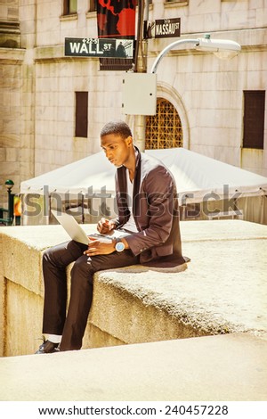 Young man working on street. A young black college student is sitting outside, looking down, working on a laptop computer. Wall Street sign in the background.