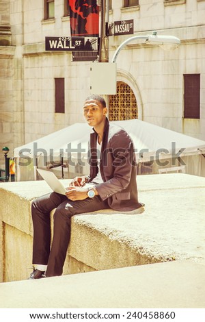 Young man working on street. A young black college student is sitting outside, looking away, narrowing eyes, thinking, working on a laptop computer. Wall Street sign in the background.