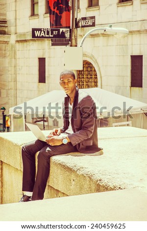 Young man working on street. A young black college student is sitting outside, looking away, thinking, working on a laptop computer. Wall Street sign in the background.