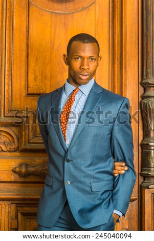Dressing formally in blue suit, patterned shirt and necktie, short haircut, a arm crossing back, a hand in pocket, a young guy is standing in the front of an old style office door, looking at you.