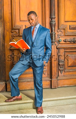 Dressing in blue suit, patterned shirt, necktie, short haircut, wearing ear stud, a young black teacher is standing in the front of old style office door, feet stepping on stairs, reading a red book.