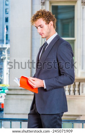 Man Reading Outside. Dressing in black suit with Shawl Lapel, black neck tie, a young sexy guy with curly hair is standing inside office building, looking down, hands opening a red book, reading.