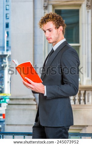 Man Reading Outside. Dressing in black suit with Shawl Lapel, black neck tie, a young sexy guy with curly hair is standing inside office building, hands holding a red book, looking down, reading.