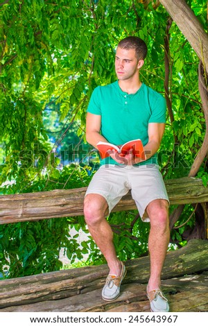 Young man reading outside. Wearing a green short sleeve Henley shirt, light yellow shorts, leather casual shoes, a young handsome college student is sitting on tree trunk in woods, reading a red book.