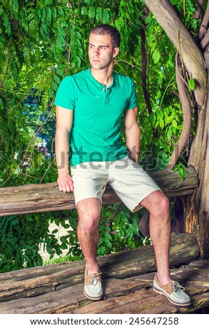 Young man relaxing outside. Wearing a green short sleeve Henley shirt, light yellow shorts, leather casual shoes, a young handsome guy is sitting on tree trunk in woods in hot summer.