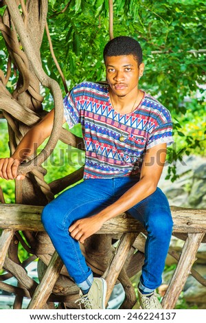 Young Black Man Relaxing Outside. Wearing a colorful pattern shirt, blue jeans, necklace, a young handsome guy is sitting on tree trunk, charmingly looking at you.