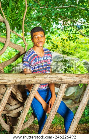 Young Black Man Waiting for You. Wearing a colorful pattern shirt, blue jeans, sneakers, necklace, a young handsome guy is sitting behind a wooden fence, charmingly looking at you.