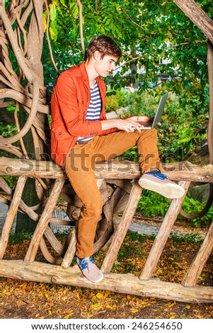 Man working outside. Wearing a dark reddish brown jacket, unbuttoned, brown corduroy pants, a young college student sitting on a wooden fence with rattan trees, reading, working on laptop computer.