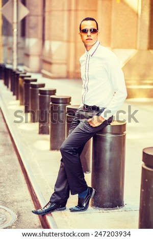 Man Street Fashion. Dressing in a white shirt, black pants, leather shoes, wearing sunglasses, a young, mysterious guy is sitting on the street, relaxing and waiting for you. Instagram filtered look.