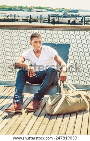 Traveling. Wearing white, V neck T shirt, blue pants, brown boot shoes, a hand carrying duffel bag, a young black college student is sitting on dock by the river, relaxing, thinking, lost in thought.