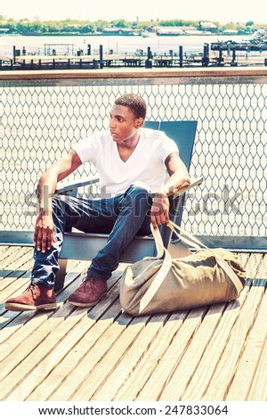 Wearing white, V neck T shirt, pants, boot shoes, a hand carrying duffel bag, a young black college student is sitting by river, thinking. Concept of young people thinking about long journey of life.