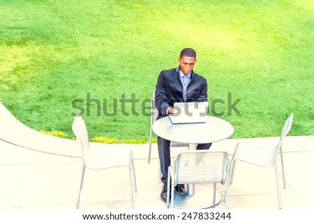 A young black college student is sitting by green lawn on campus, looking down, working on a laptop computer, connected with wireless Internet. Concept of modern life, technology and environment.