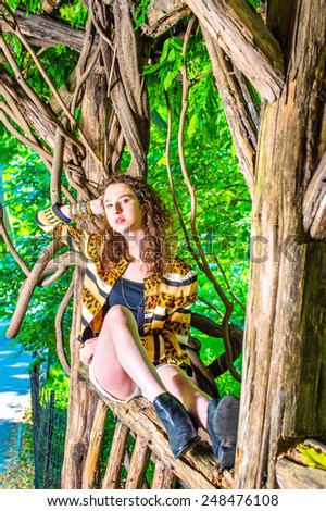 A teenager girl with curly hair sitting on tree trunk, arm resting on a rattan, supporting head, relaxing, thinking. Relaxing at cool place in hot summer. Girl patiently waiting for you on park.