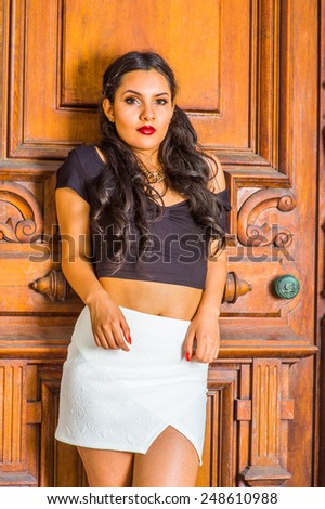 Girl waiting for you. Dressing in a black, short sleeve top, white short fit skirt, a young pretty lady with long curly hair is standing by an old fashion style door, charmingly looking at you.