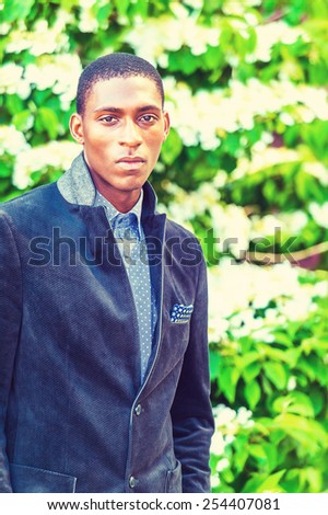Portrait of young teenage boy. Dressing in a fashionable jacket, a young black college student is standing outside by green plants, white flowers, innocently looking at you. Retro filtered look.