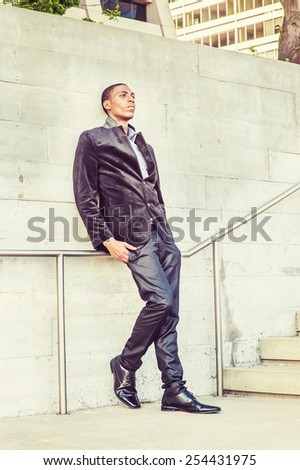 City Life. Wearing a fashionable jacket, pants, leather shoes, a young black college student standing against the wall, sad, thinking. Concept of teenager self esteem. Retro filtered look.