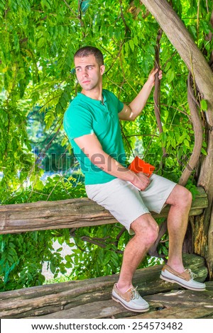 Wondering. Wearing short sleeve shirt, shorts, leather casual shoes, holding red book, a young guy sitting on tree trunk, looking around. Concept of introspection, self questioning, examination.
