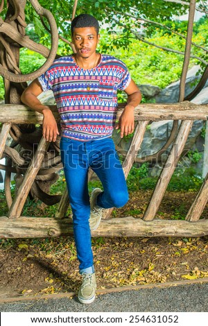 Young Black Man Waiting for You. Wearing a colorful pattern shirt, blue jeans, sneakers, necklace, a young handsome guy is standing against a wooden fence, charmingly looking at you.