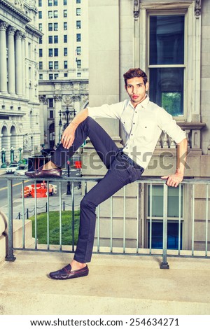 Man Casual Fashion. Wearing a white shirt, black pants, leather shoes, a young college student is sitting on a railing in a old fashion street, bending a leg, relaxing. Instagram filtered look