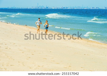 Young couple running on Sandy Hook Beach, New Jersey, guy wearing gray t shirt, yellow pants, girl dressing in strapless blue sun dress. Far background is busy New York City. Instagram filtered look.