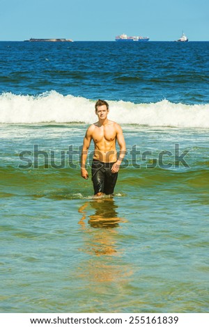 Portrait of Young Sexy Man. Well built young handsome muscular guy, half naked, wearing bathing suit, walking out of water on the beach, narrowing eyes under strong sun shine. Instagram filtered look.