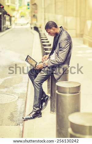 Working anywhere, anytime. Young man working on street. A young black college student is sitting outside an office building, typing on a laptop computer, reading, thinking. Retro filtered look.